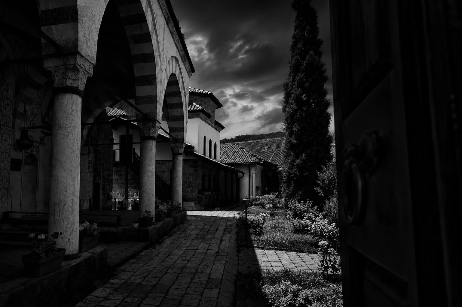 The Hadum Mosque's arcaded courtyard — striped stone arches, a cypress and a cobbled path under a dramatic sky.